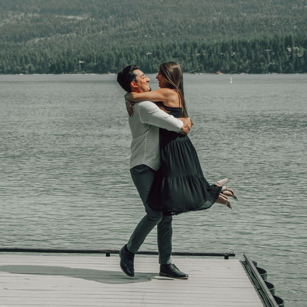 Couple embracing on a lakeside dock with forested mountains in the background.