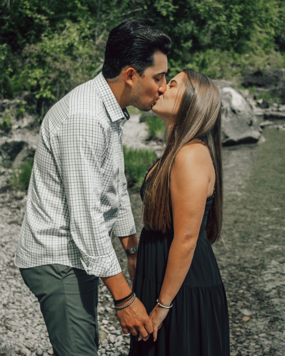 Couple sharing a romantic kiss while holding hands near a rocky creek surrounded by lush green trees.