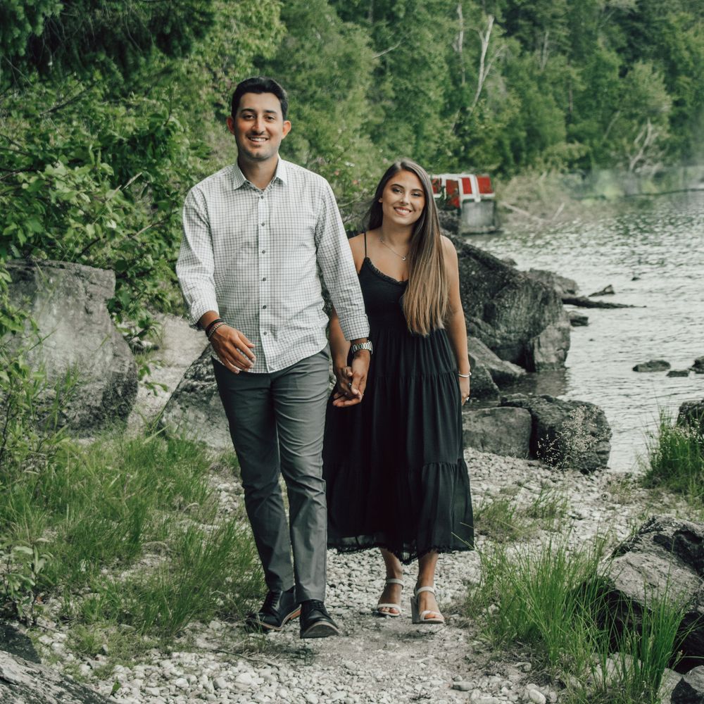 Couple holding hands and smiling while walking along a rocky lakeside path surrounded by lush green trees.