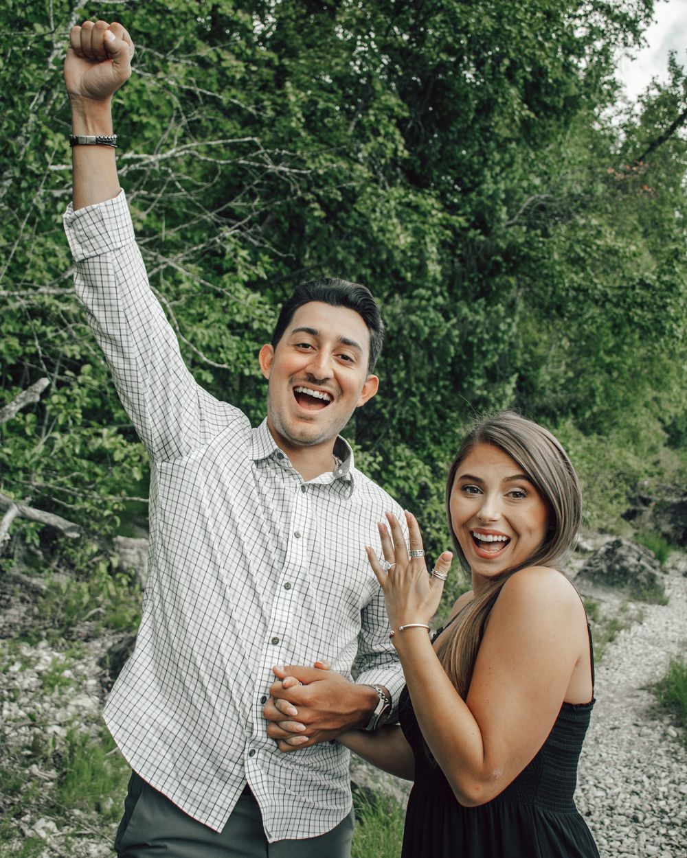 Excited couple celebrating engagement outdoors, man raising fist while woman shows off her new ring.