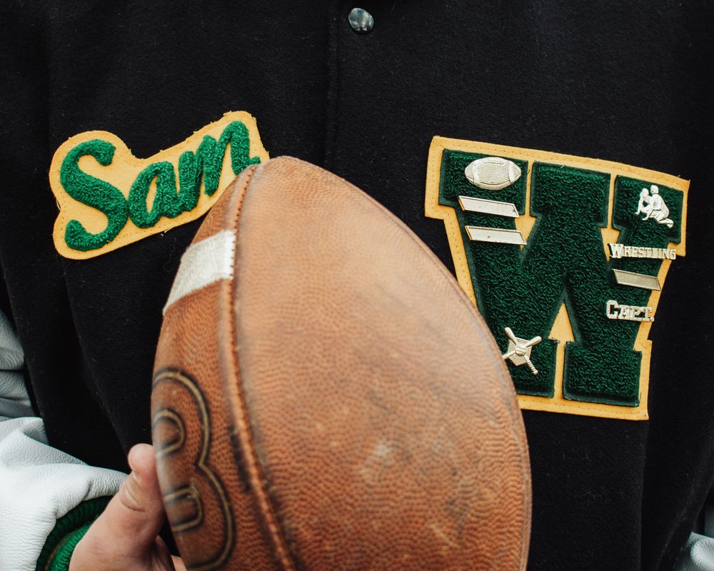 A football spirals through the air against a dark background with green and gold sports banners visible.