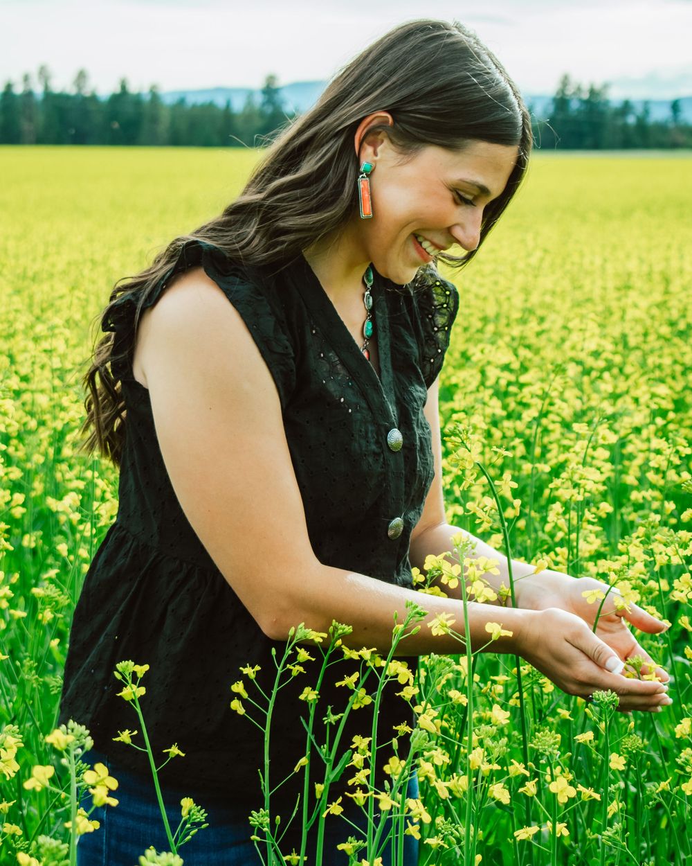 Person in black dress gently touching yellow canola flowers in a vast blooming field on a sunny day.