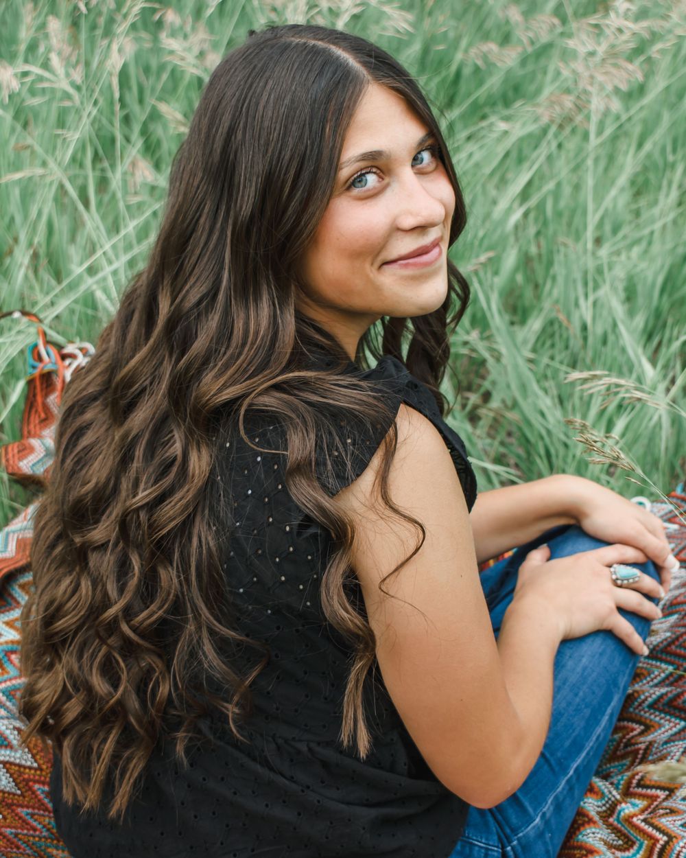 Young woman in black top and jeans sits in tall grass field while smiling and looking away from camera.