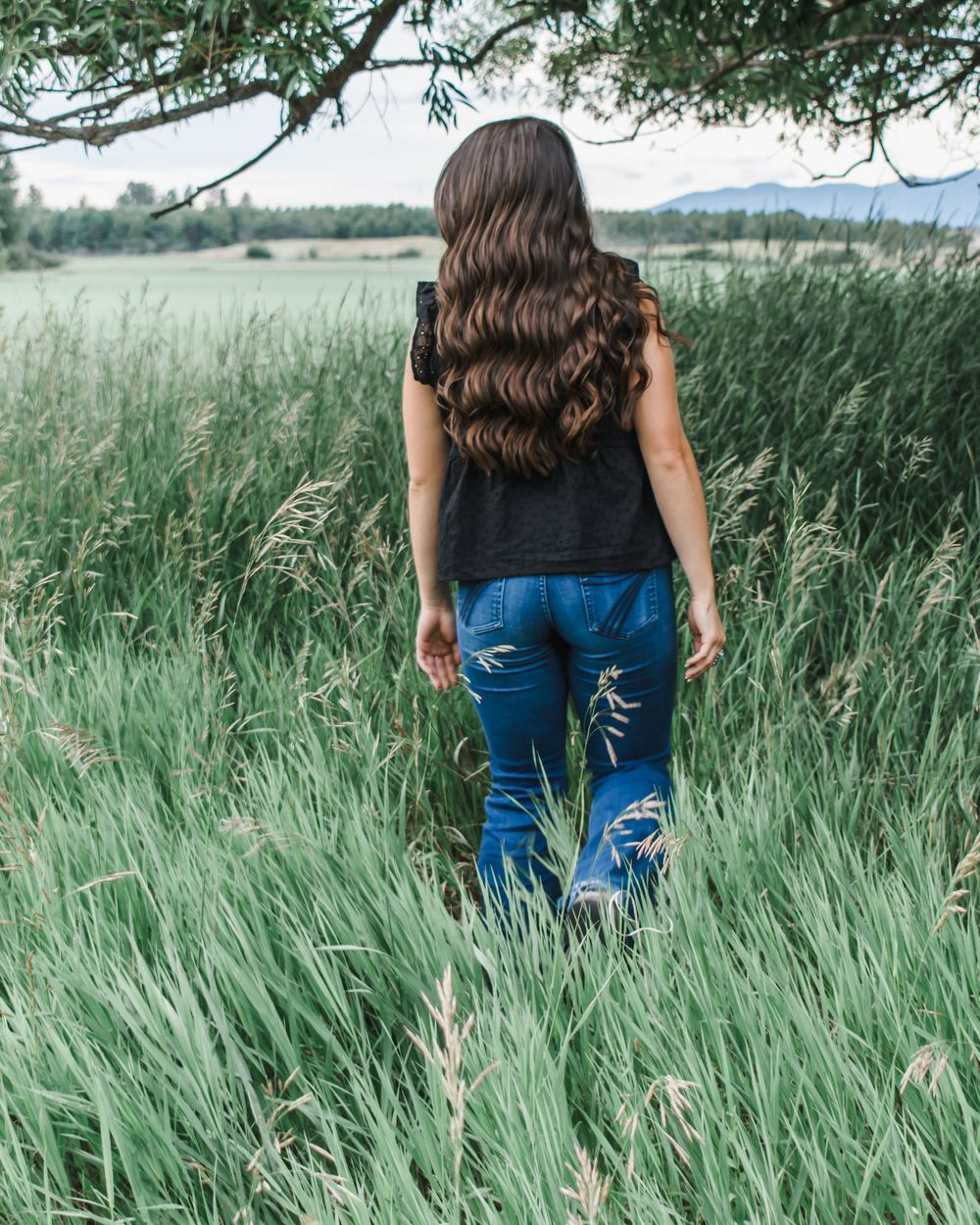 Woman in blue jeans stands among tall green grass in a serene outdoor field setting.