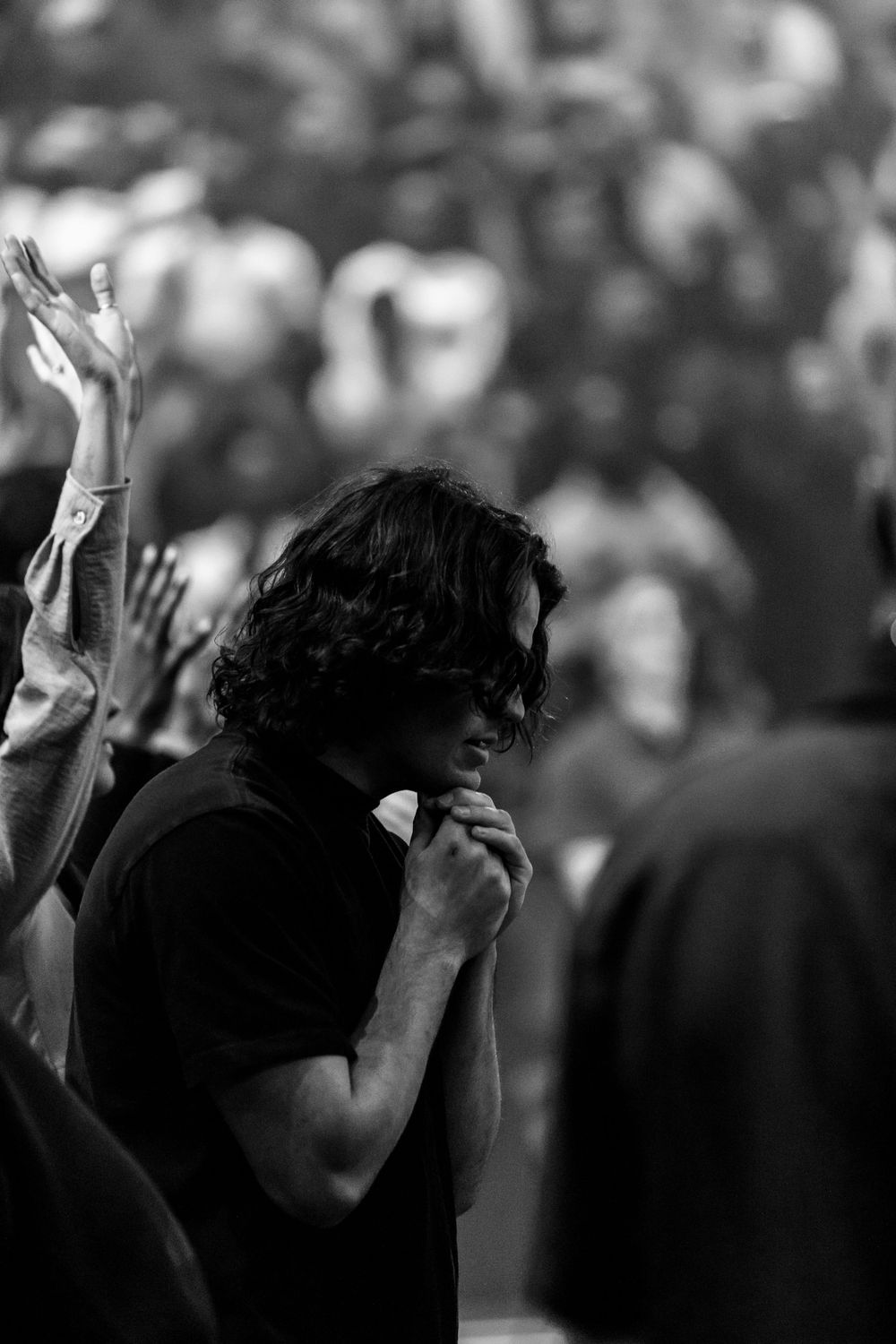 A man with curly hair prays with clasped hands amid a crowd of worshippers in black and white.