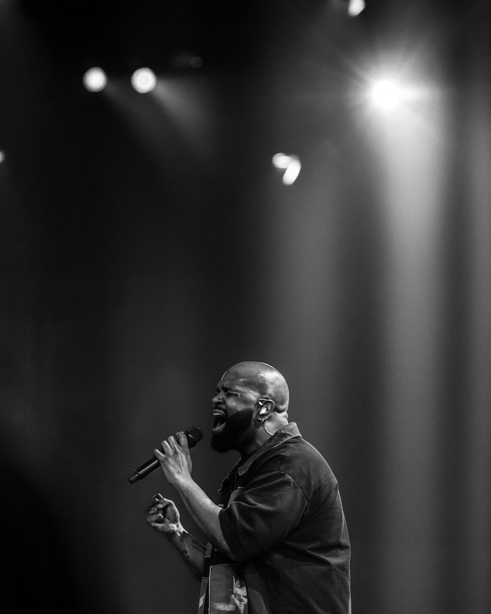 A bald man sings passionately into a microphone under dramatic stage lighting in black and white.