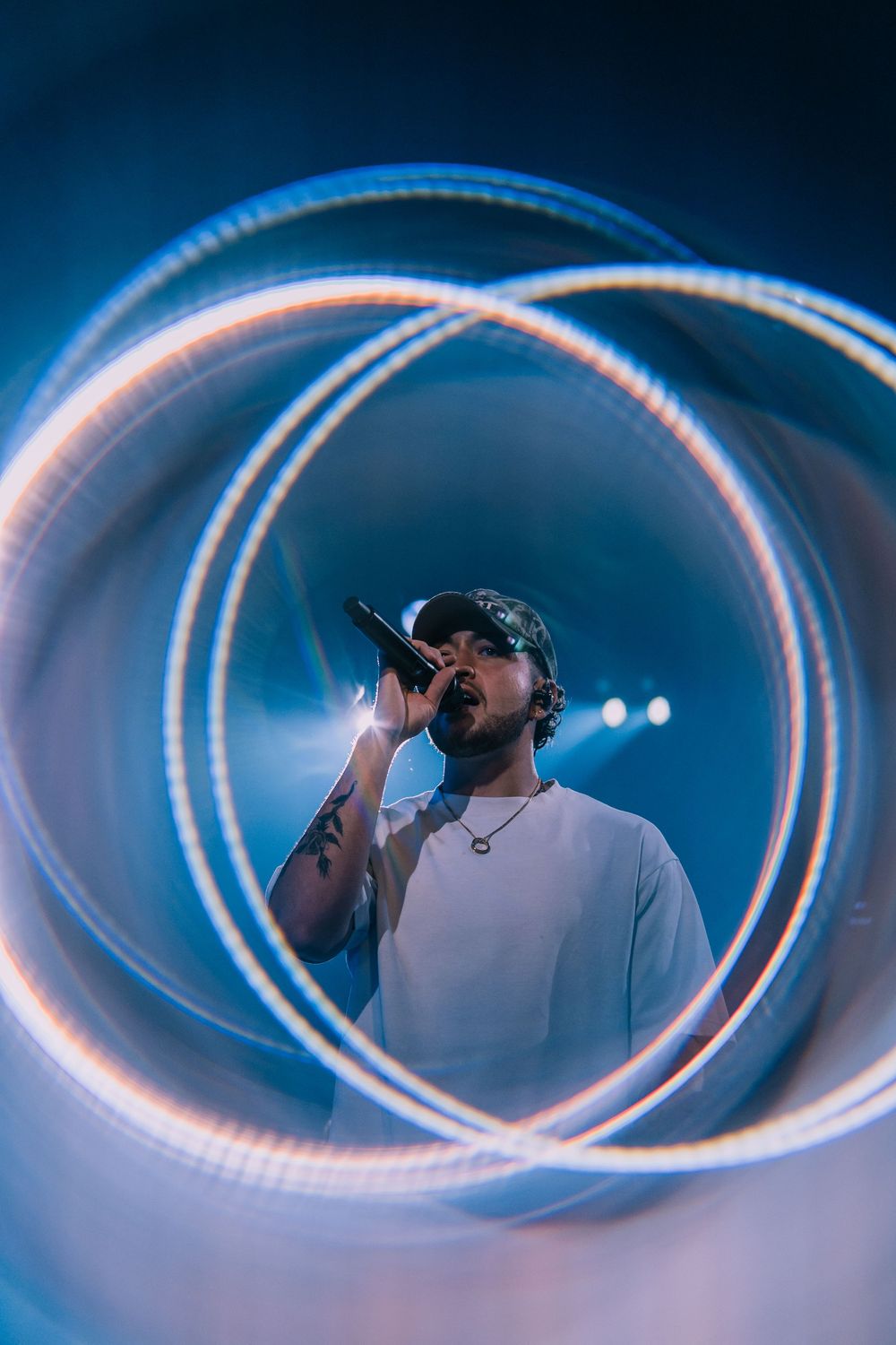 Tattooed performer singing into microphone surrounded by swirling light trails in blue-lit venue.
