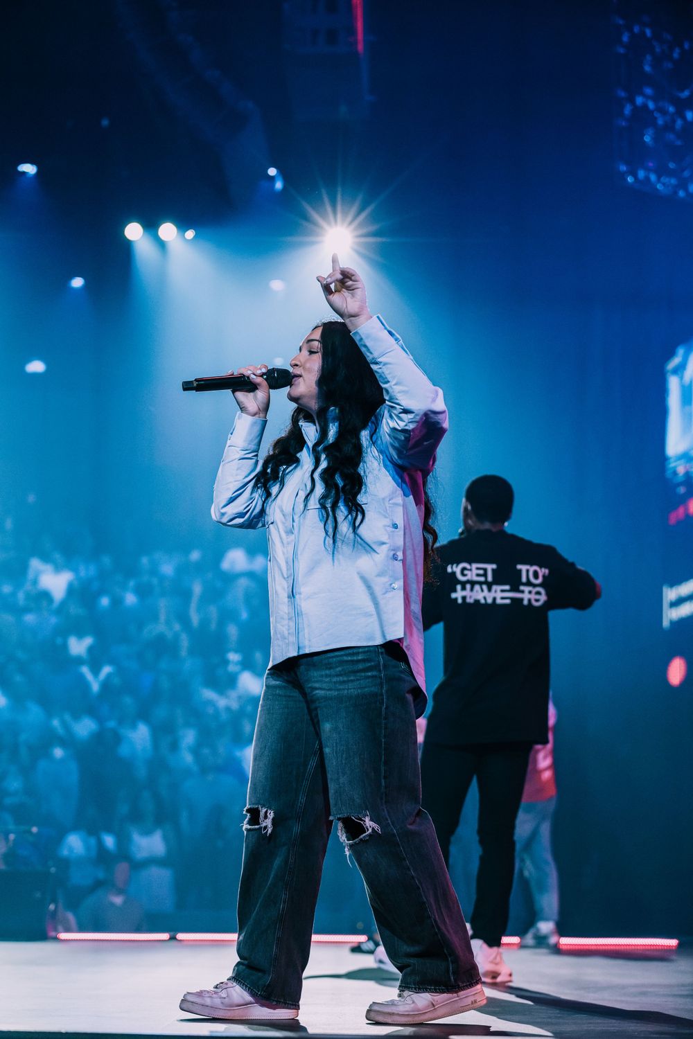 A female singer performs on a blue-lit stage with one arm raised holding a microphone.