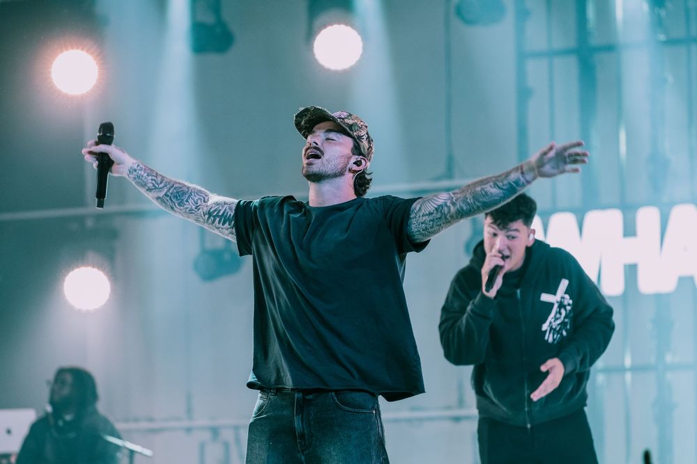 Tattooed male singer performing on stage with arms outstretched holding a microphone.