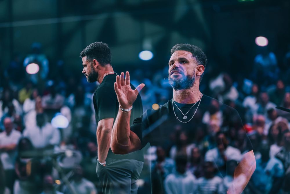 Man in black shirt raising hand in prayer at a large worship gathering event.