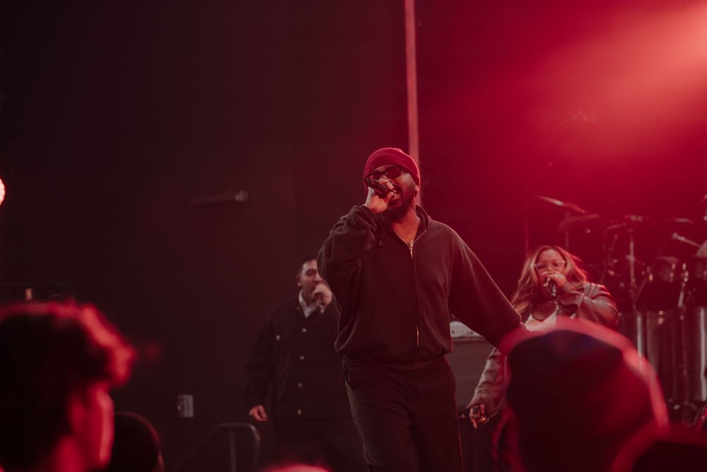 Male performer in red beanie sings into microphone on stage bathed in dramatic red concert lighting.