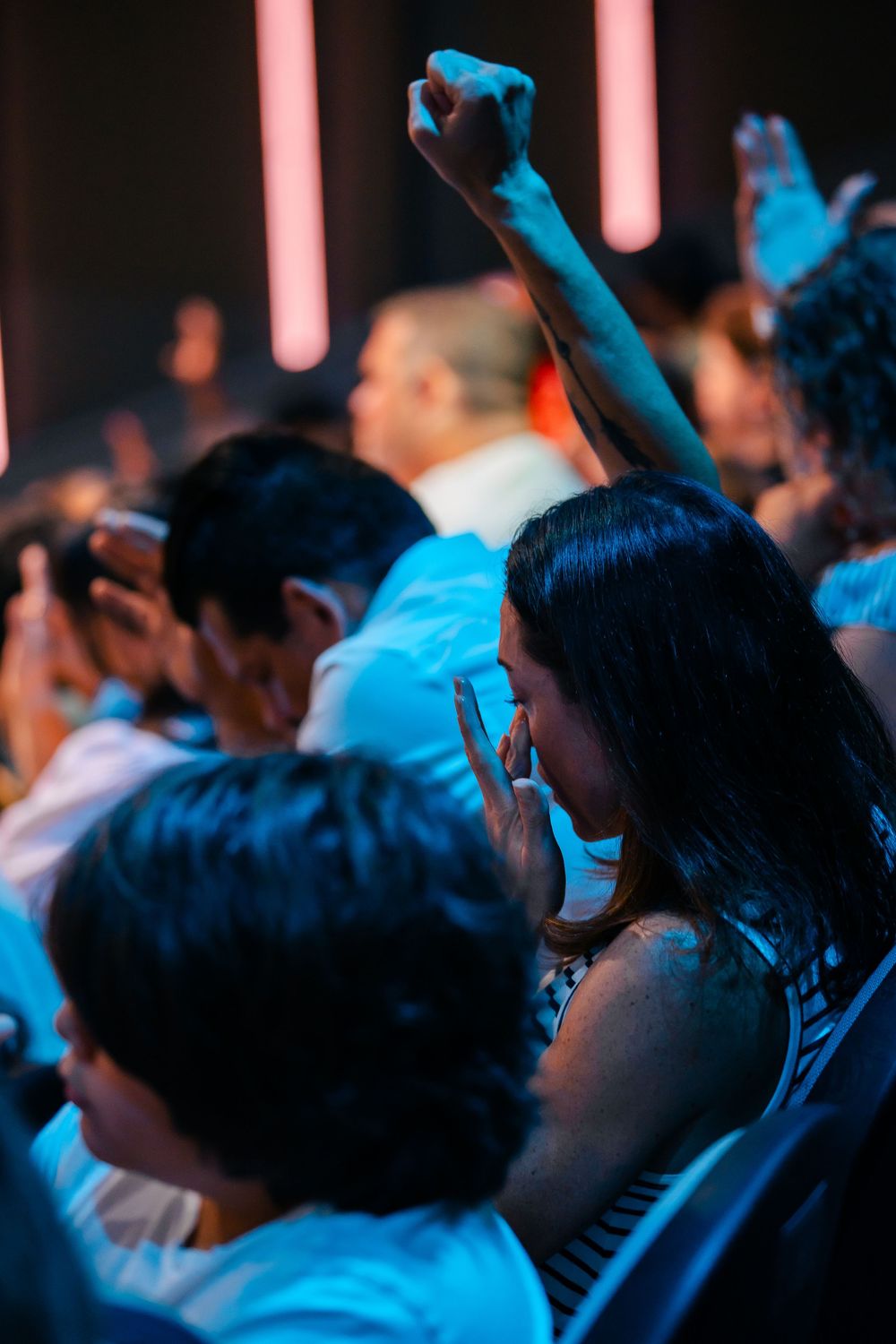 A woman raises her fist in worship at a crowded concert or church service, bathed in blue and red stage lighting.