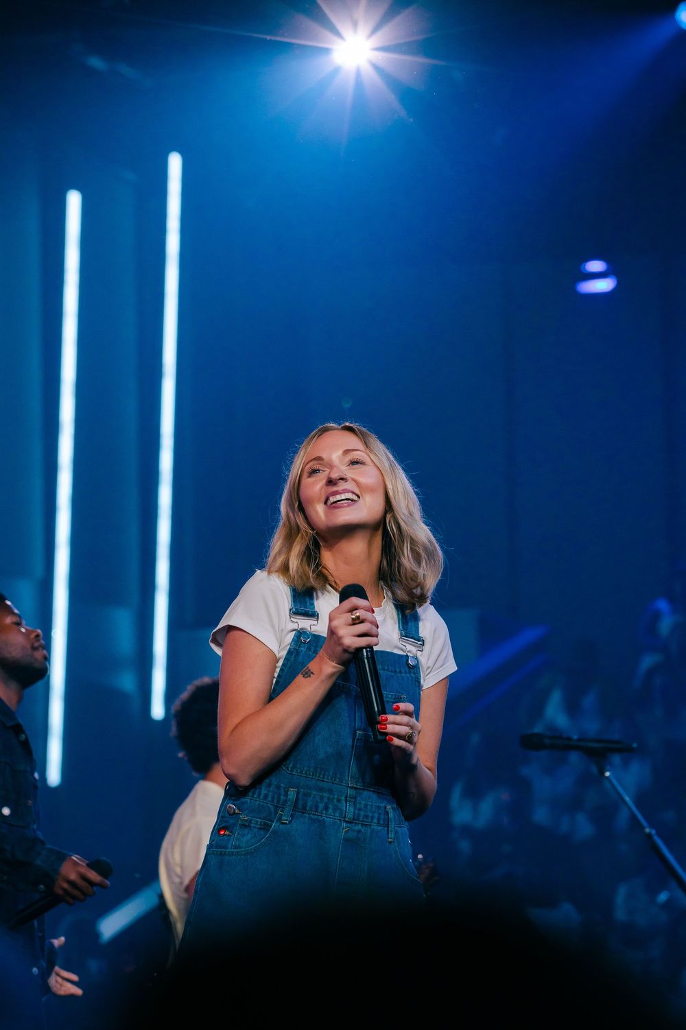 Young woman in denim overalls smiling on stage holding microphone under blue concert lights.