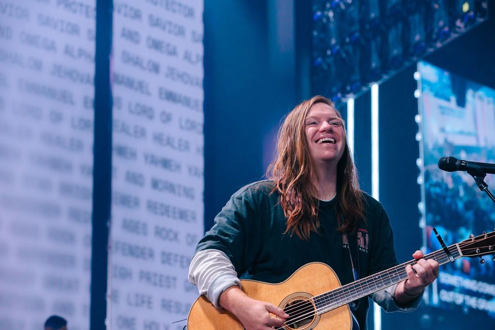 Smiling musician with long hair playing acoustic guitar on stage with colorful word backdrop.