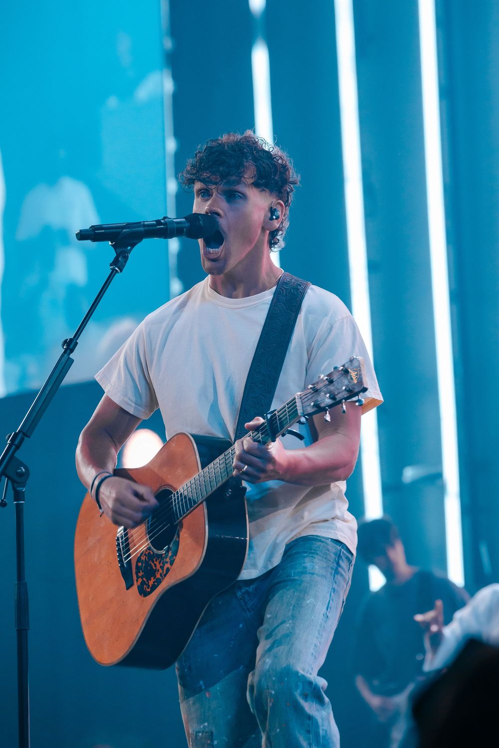 Young male musician playing acoustic guitar and singing passionately on stage with blue neon lights backdrop.