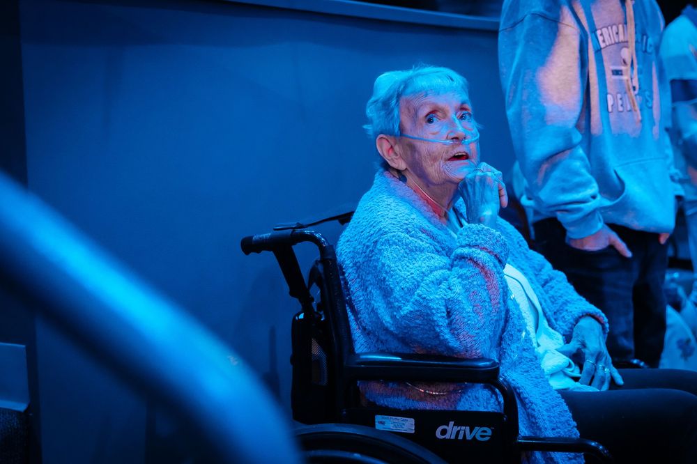 Elderly woman in wheelchair with oxygen tubes, wearing blue robe under blue lighting at an indoor event.