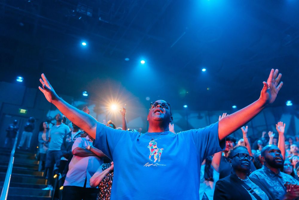 Man in blue t-shirt raises hands in worship at a vibrant church concert with blue stage lighting.