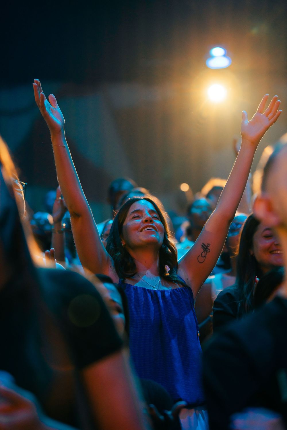 Young woman in blue dress raises arms joyfully in a crowd at a concert under warm stage lighting.