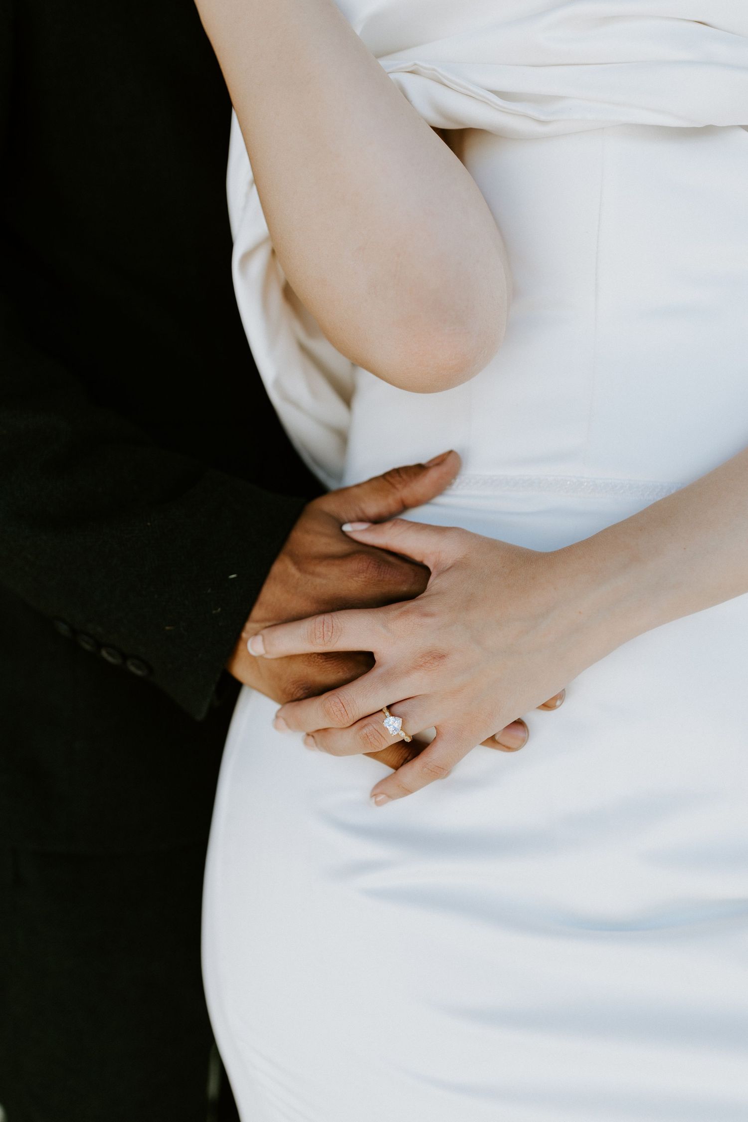 Close-up of hands with engagement ring resting on a white dress against dark background.