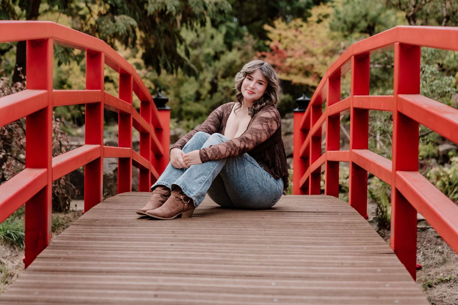 Young woman sitting on a red wooden bridge surrounded by autumn foliage in a serene garden setting.
