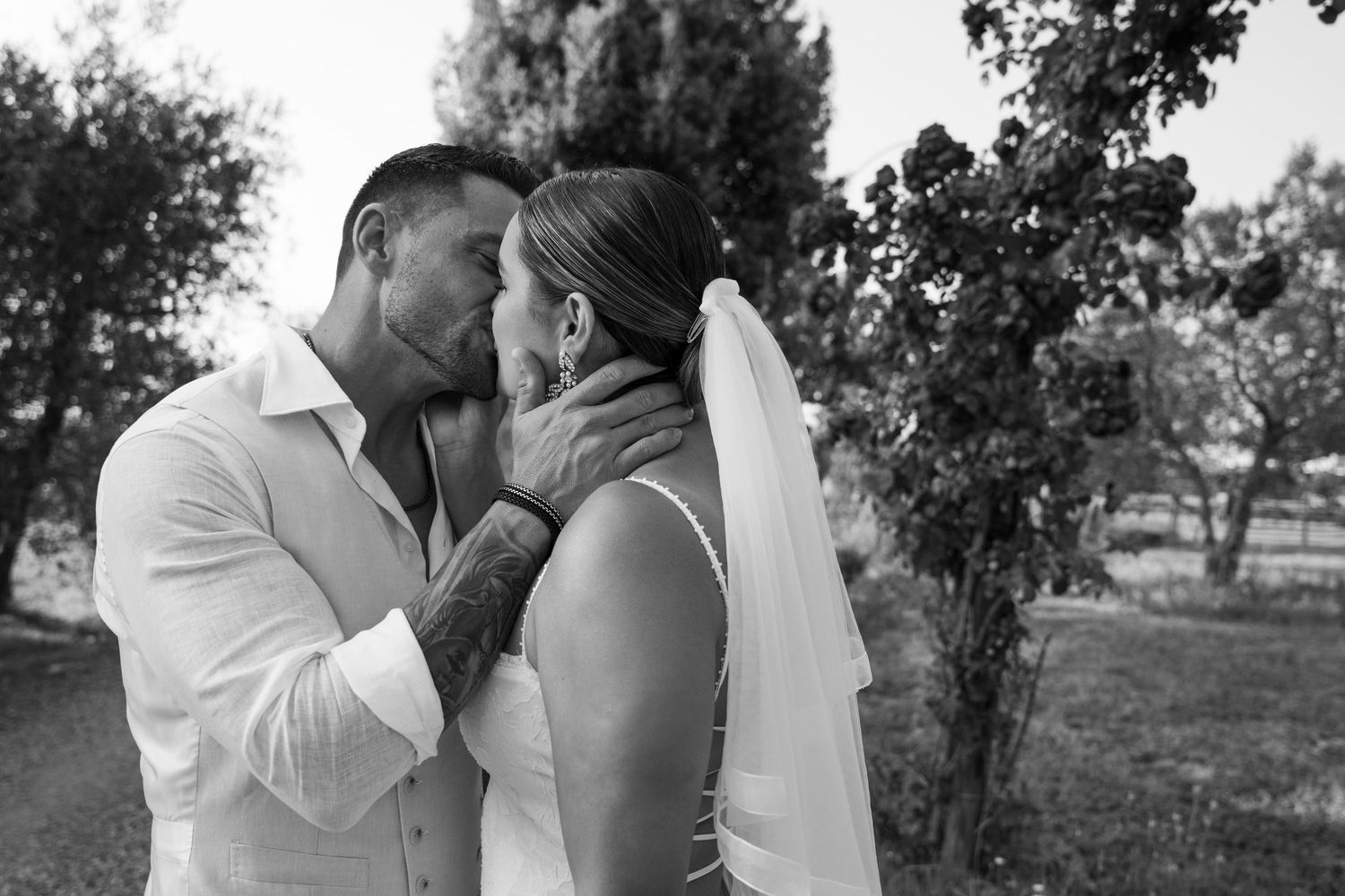 Black and white candid of a bride and groom kissing in an outdoor, tree-lined setting; authentic destination wedding and couple portraiture.