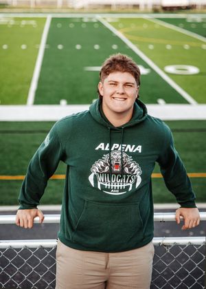 Young man in Alpena Wildcats green hoodie smiling on football field bleachers.
