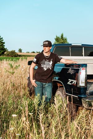 Young man in black graphic tee and snapback hat leaning on a blue Chevy Z71 truck in a rural field.