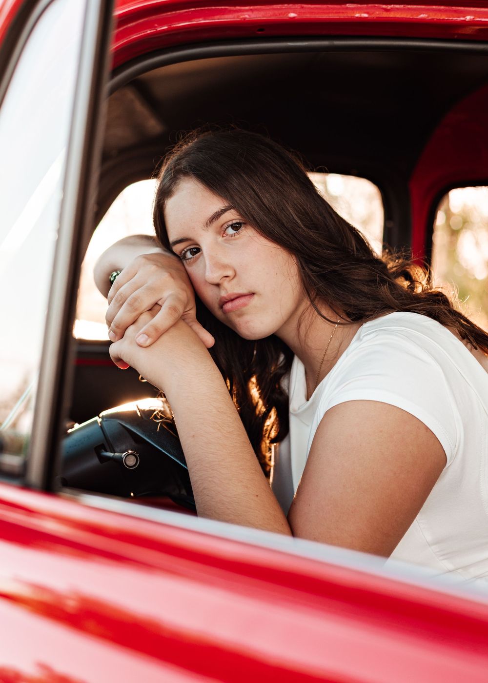 Young woman with dark hair rests her chin on hands over steering wheel inside a vintage red car.