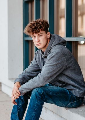 Young person in gray hoodie and jeans sits casually on concrete ledge near modern building.