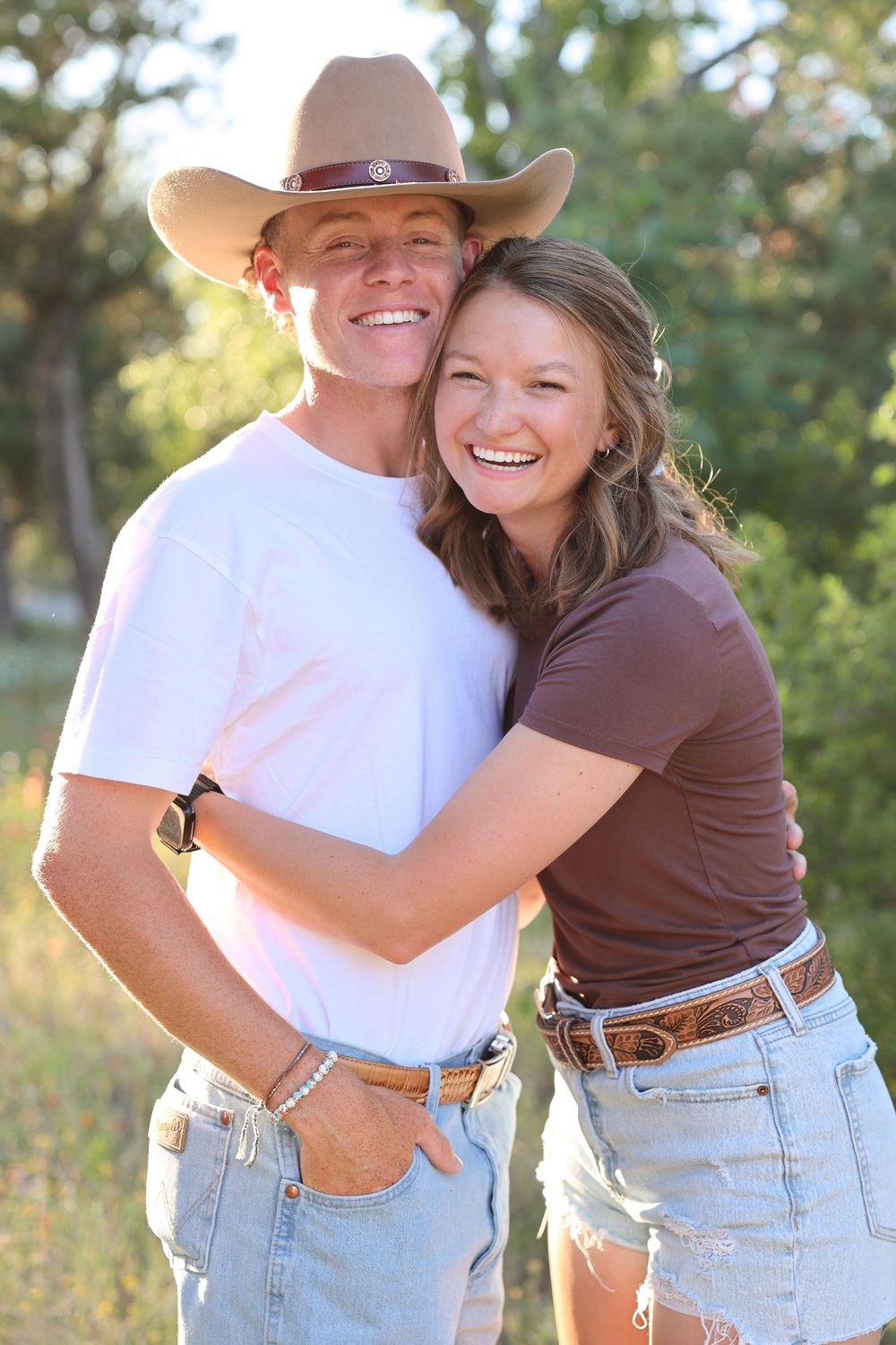 A couple in casual western attire smile together outdoors against a sunlit natural background.