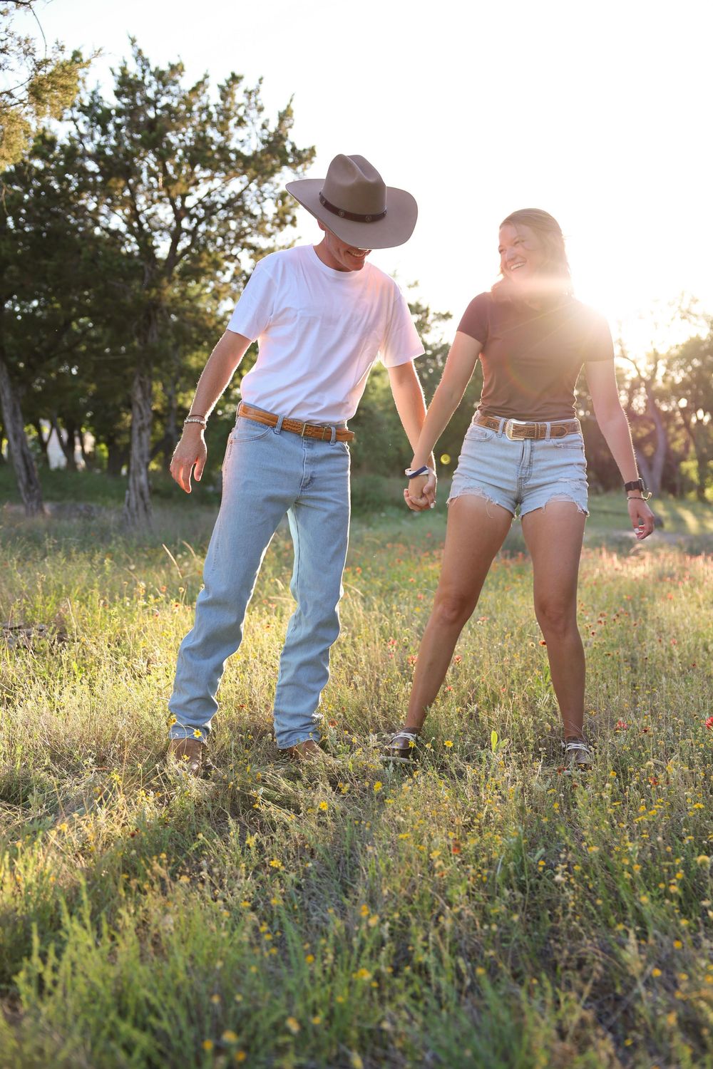 A couple holds hands while standing in a sunlit meadow at sunset wearing casual western-inspired outfits.