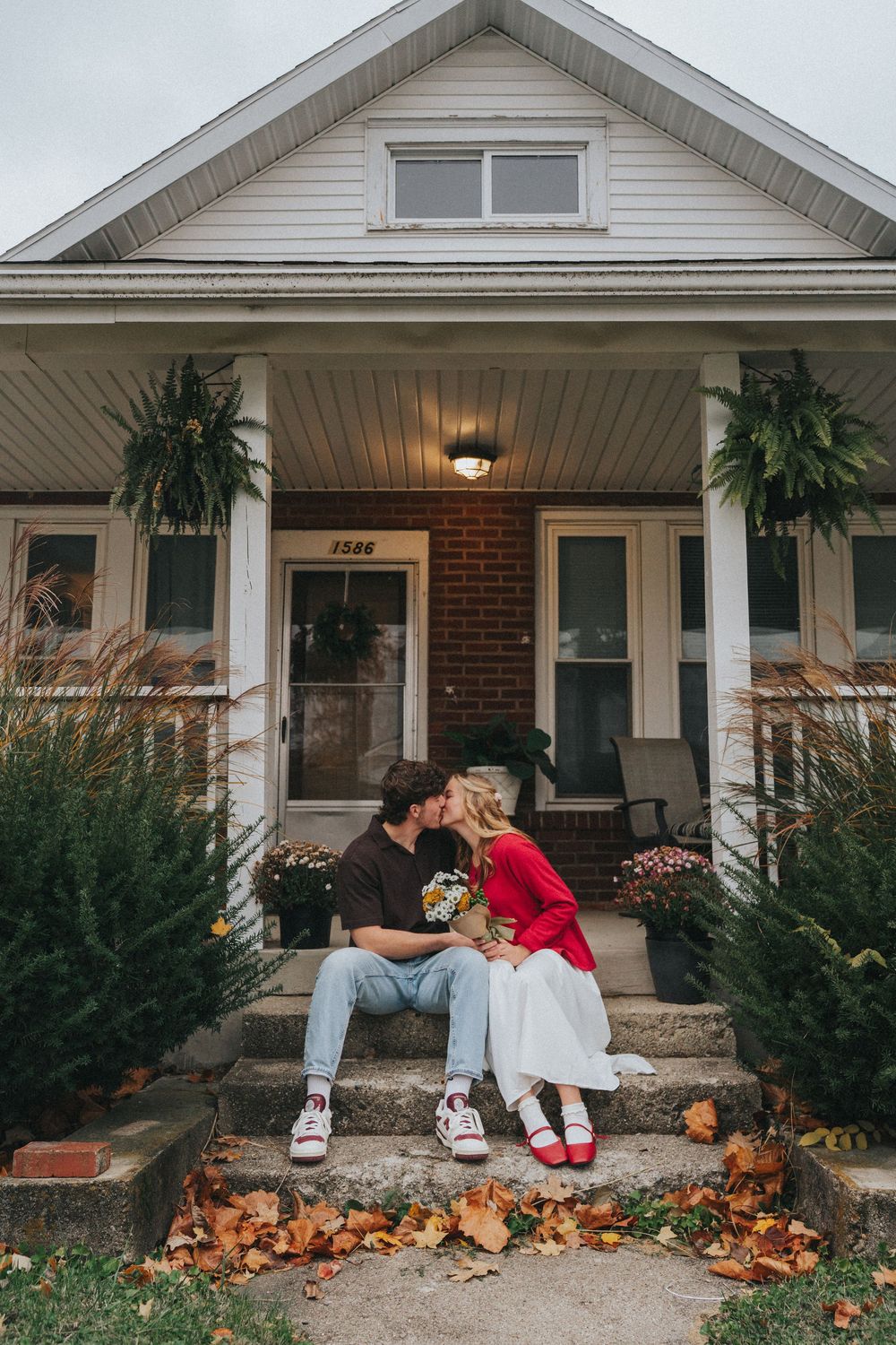 Couple kissing on front porch steps of white house in autumn, woman in red jacket holding bouquet.