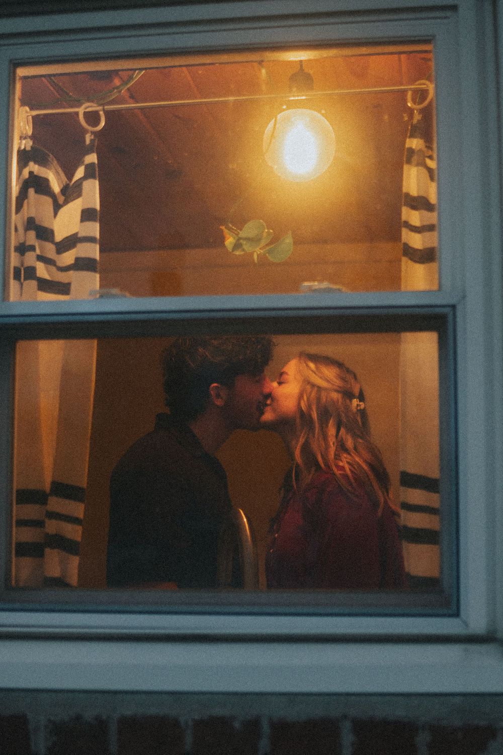 Couple sharing a romantic kiss framed in a warmly lit window at dusk, with striped curtains visible inside.