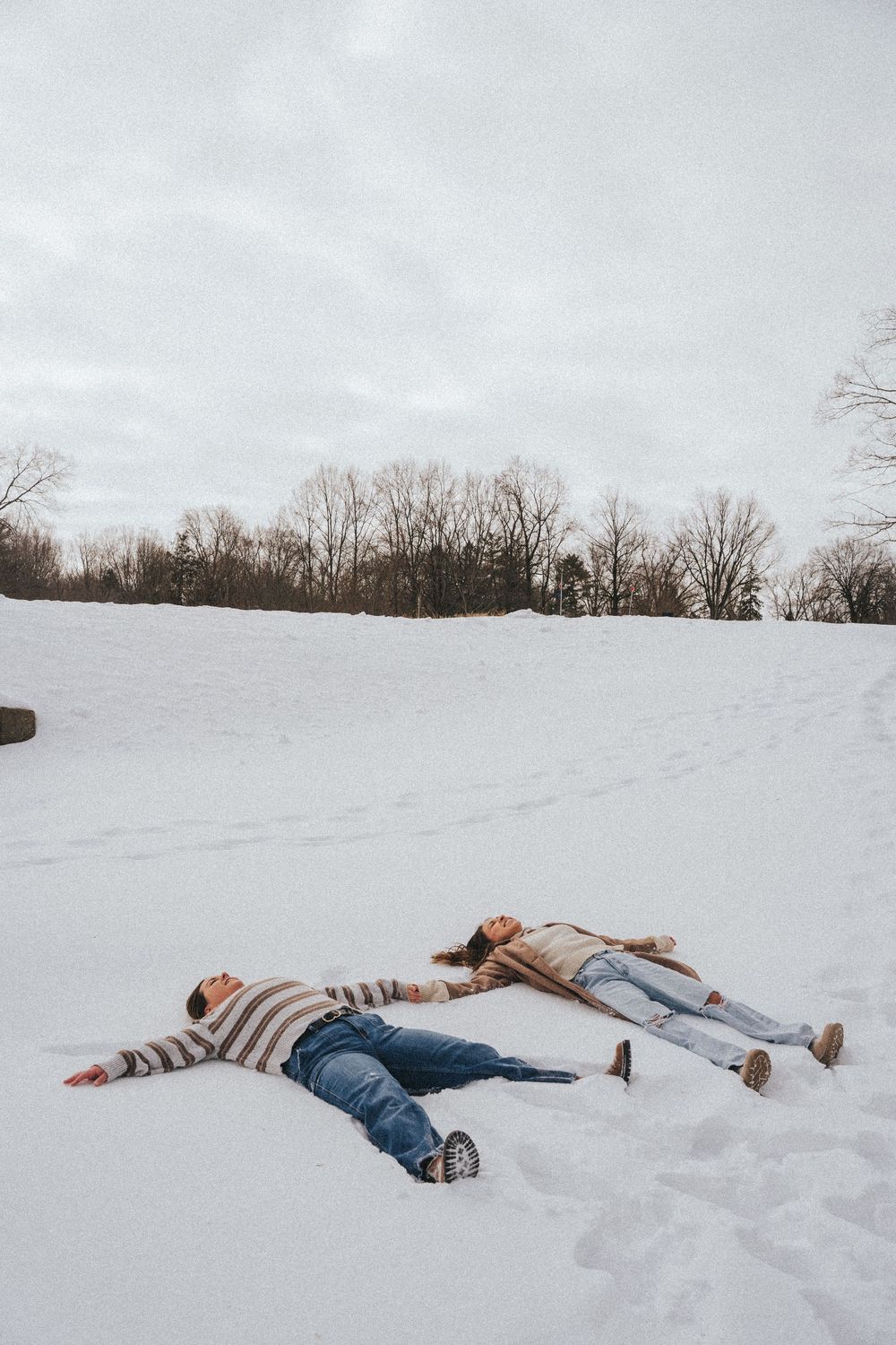 Couple making snow angels on a snowy field with bare trees in the background on a cloudy winter day.