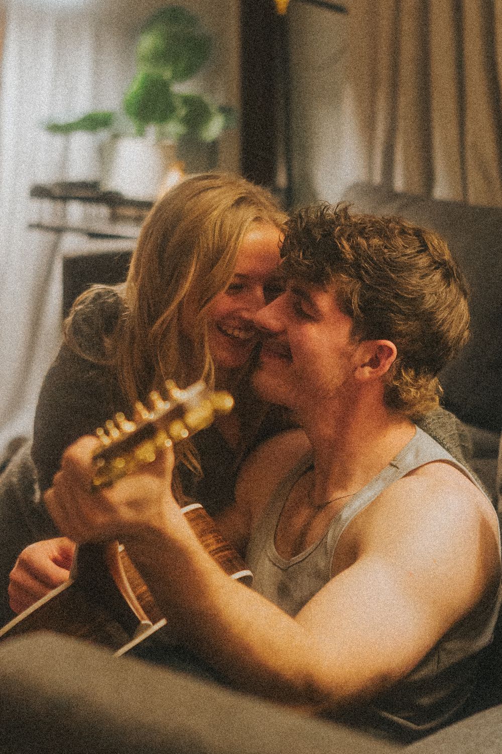 A young couple sharing a tender moment while playing acoustic guitar together on a cozy couch indoors.