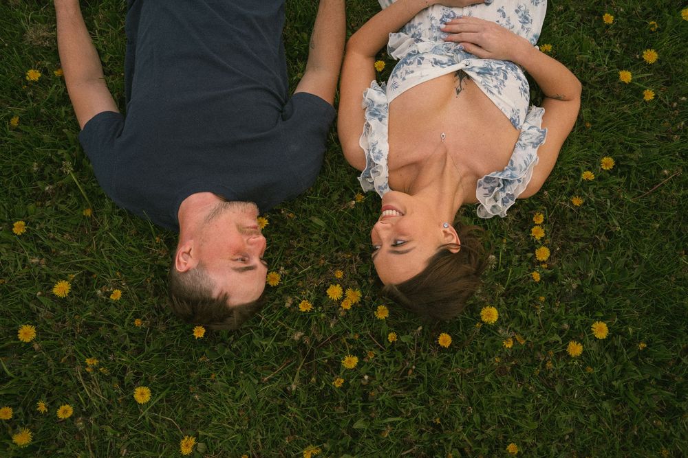 Couple lying on green grass with yellow wildflowers, smiling at each other in a romantic outdoor setting.