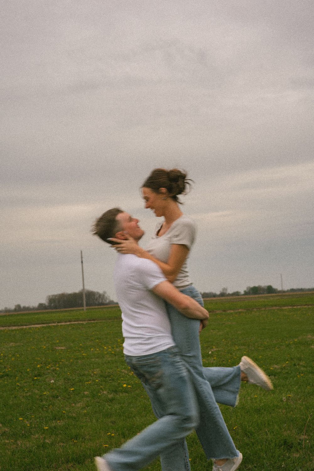 A joyful couple embraces in a green field, man lifting woman as they laugh together on a cloudy day.