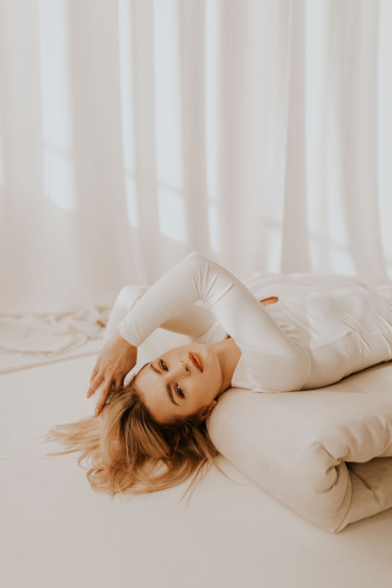 Woman in cream colored outfit reclining elegantly against white curtained backdrop.