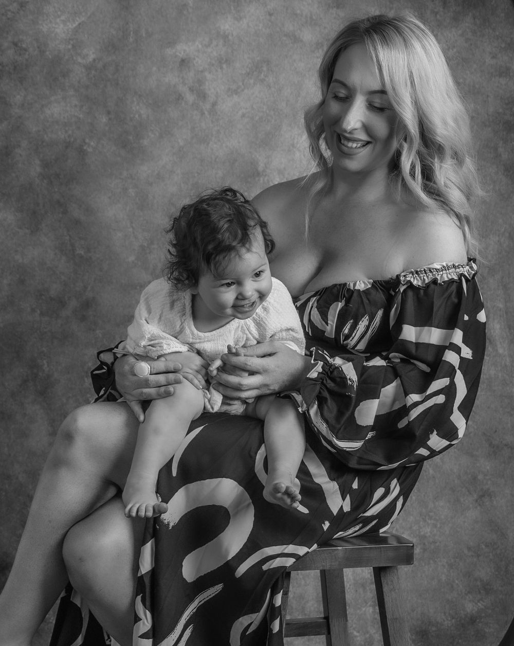 Black and white studio portrait of mother and baby bonding on wooden furniture.
