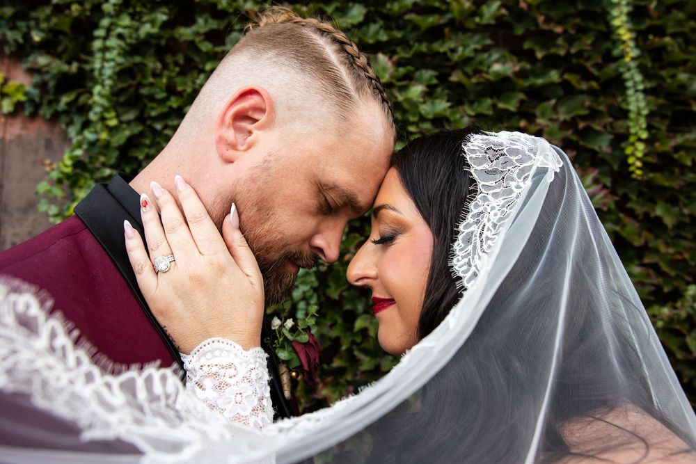 Bride and groom touch foreheads in a tender wedding moment, her lace veil draped over them, with an ivy-covered wall behind.