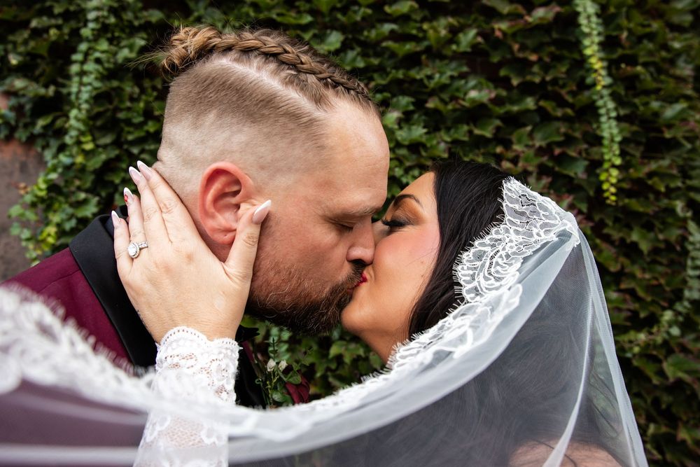 Bride in a lace veil and groom with a braided hairstyle share a romantic kiss in front of a green ivy wall on their wedding day.