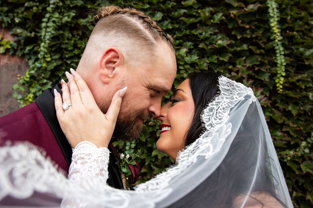 Bride in a lace veil and groom in a burgundy jacket touch foreheads and smile in a tender wedding moment against an ivy-covered wall.