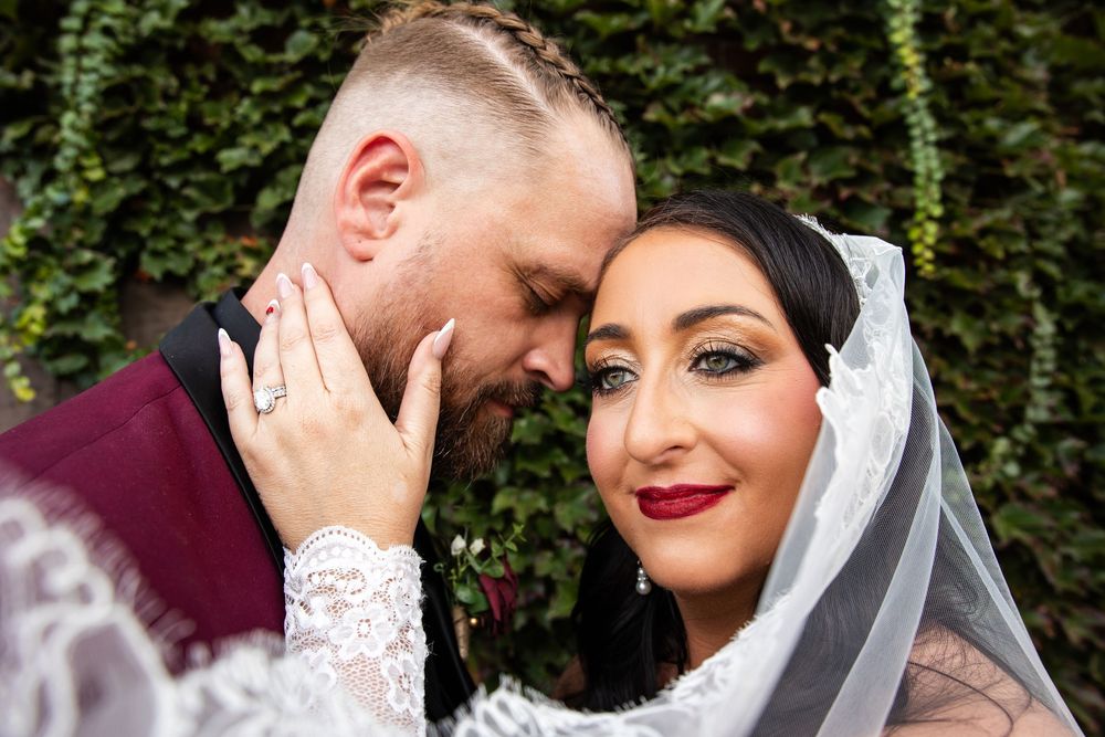 Close-up of a bride and groom embracing tenderly, the bride in a lace veil and red lipstick showing her wedding ring against ivy.