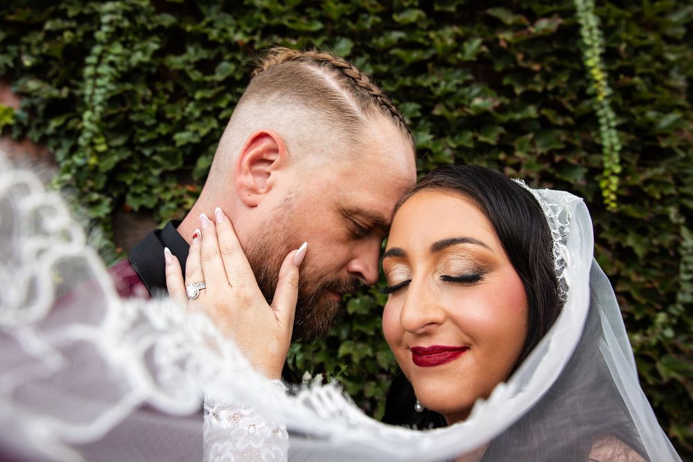 Bride in a lace veil closes her eyes and touches her groom's face as they embrace in a tender wedding portrait before a leafy green wall.