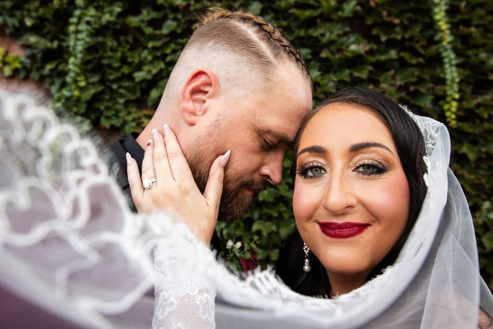 Bride with red lipstick smiles beneath a lace veil as her groom nuzzles her cheek, her hand on his face, against a green ivy wall.