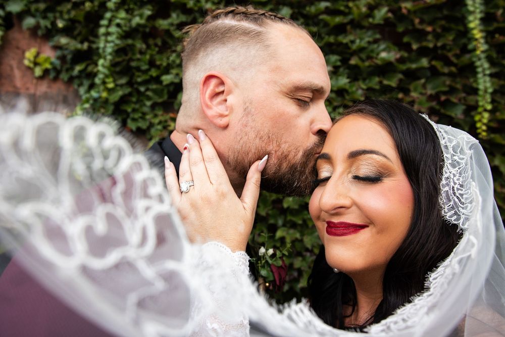 Groom kisses the cheek of a smiling bride in a lace veil as she shows her wedding ring, with green ivy in the background.