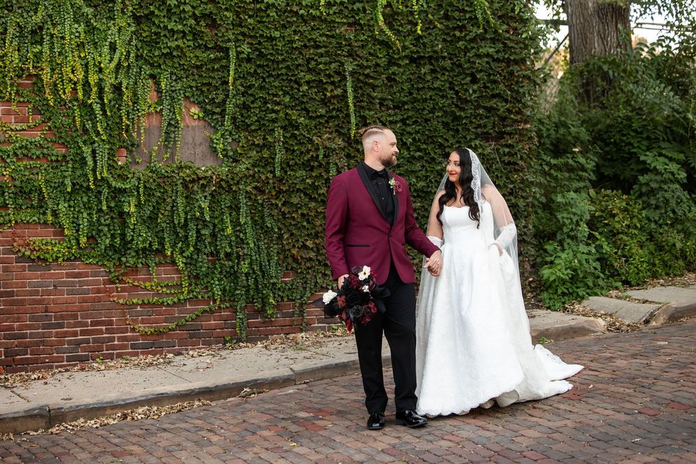 Bride in a white gown and veil holds hands with a groom in a burgundy suit on a brick street lined with ivy-covered walls.