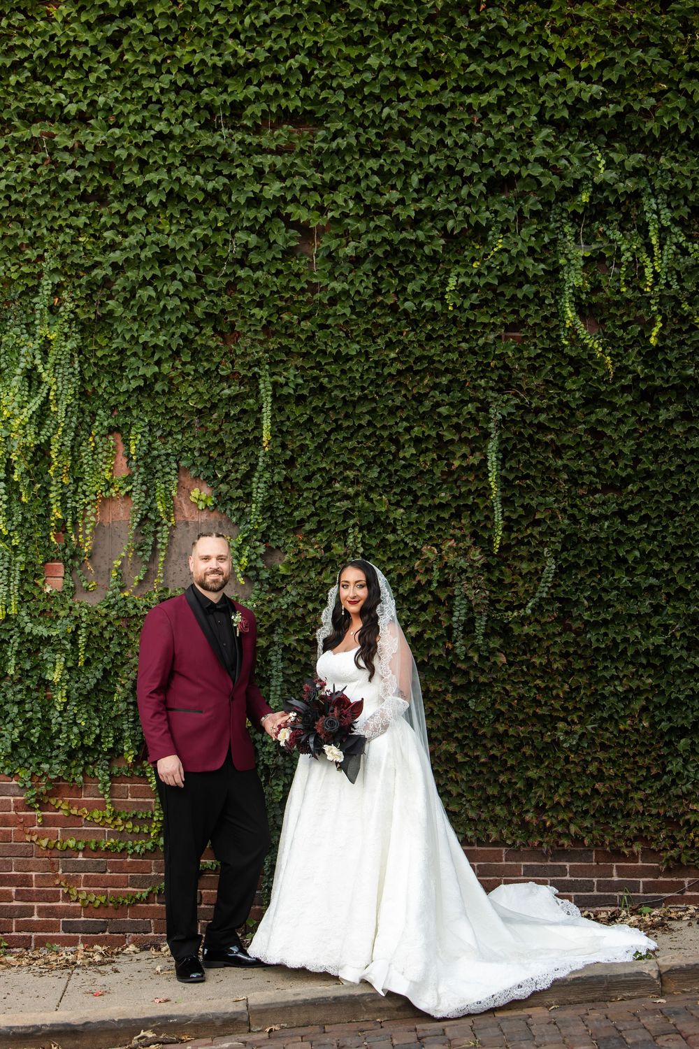 Bride in a white lace gown and veil holds a dark floral bouquet beside a groom in a burgundy blazer in front of an ivy-covered brick wall.