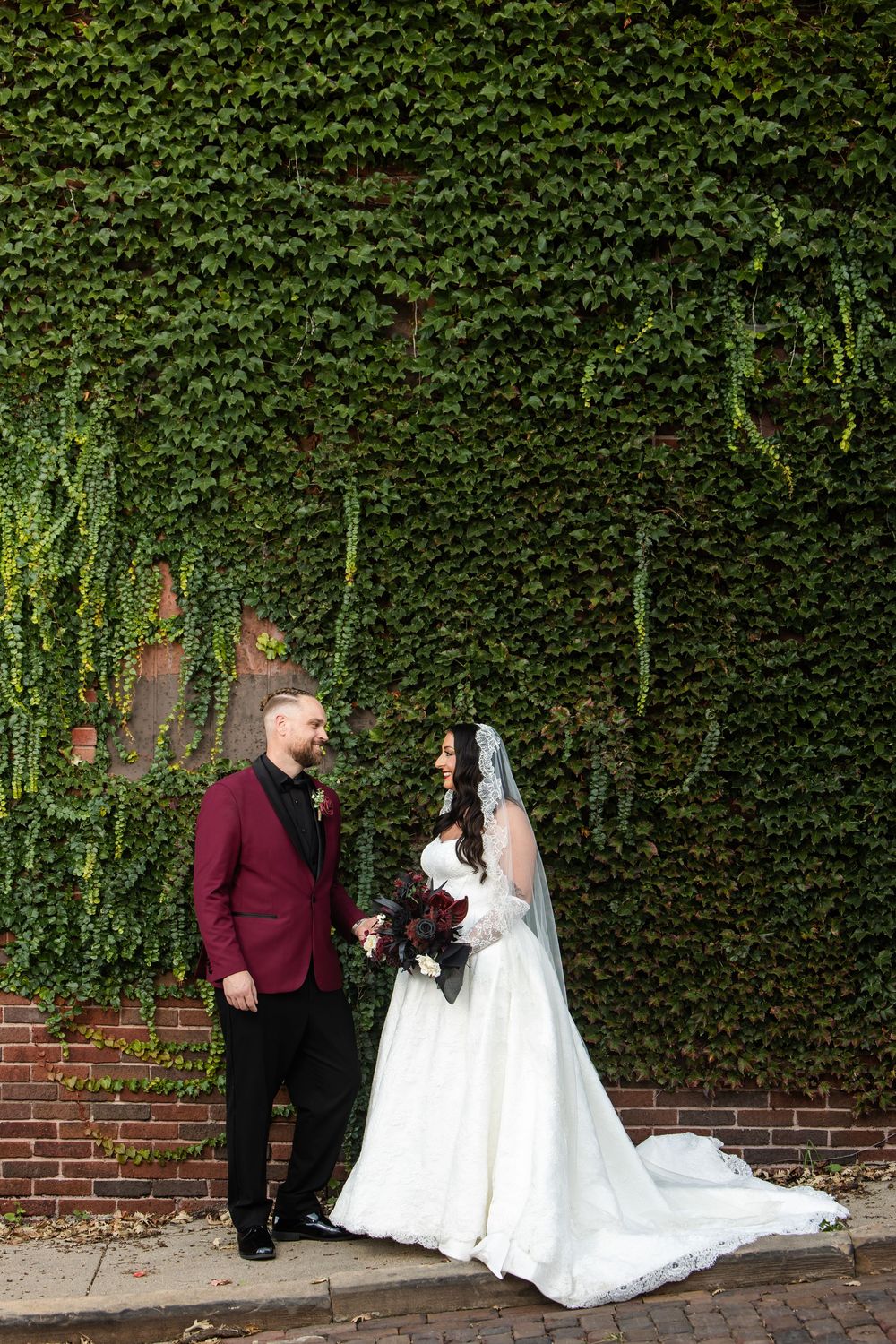 Bride in a white lace gown and veil holds hands with a groom in a burgundy blazer before an ivy-covered brick wall, bouquet in hand.