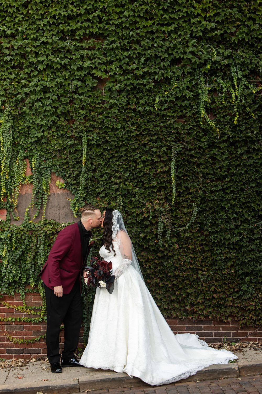 Bride in a white gown with veil and groom in a burgundy blazer share a kiss before an ivy-covered brick wall on their wedding day.